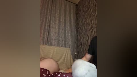 Video of a baby lying on a beige blanket, wearing a white onesie with a pink polka-dot pattern. The background features a textured, brown curtain.