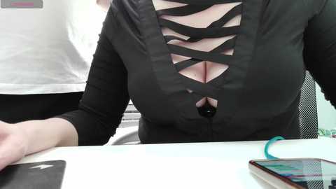 Video of a woman with a light complexion wearing a black, crisscrossed lace-up top revealing cleavage, seated at a white desk. A smartphone with colorful screen and a pen lie nearby.