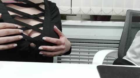 Video of a woman with fair skin wearing a black, crisscrossed lace top revealing cleavage, standing in an office with white blinds and a mesh chair.