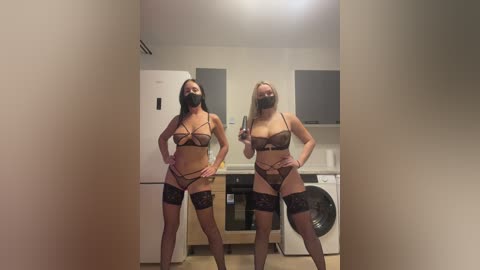 Video of two women in matching black lingerie, strappy bras, and lace thigh-highs, standing confidently in a modern kitchen with white cabinets and a white refrigerator.