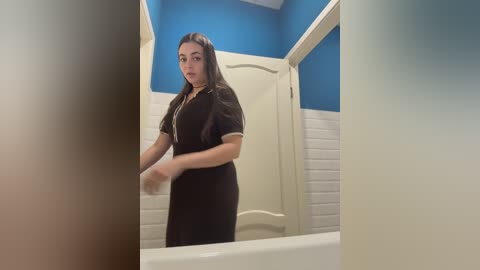 Video of a young woman with long dark hair, wearing a black dress, standing in a bathroom with white tiles and a blue accent wall. She is looking at a frosted glass door.