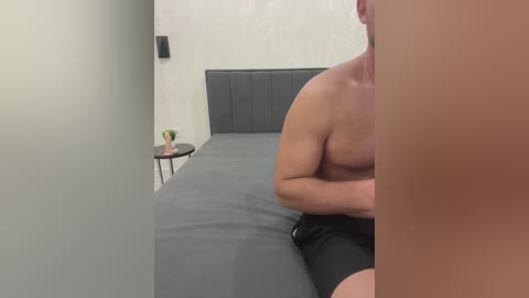 Video of a muscular, shirtless man with fair skin, wearing black shorts, sitting on a gray bed in a minimalistic room with a beige wall and a small, round side table with a vase.