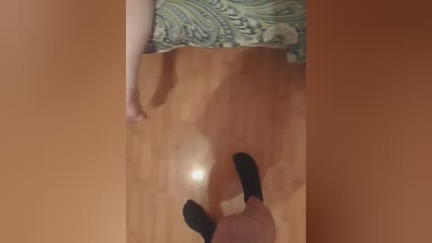 A video of a woman's legs and feet in black socks, standing on a wooden floor. She wears a green and blue paisley-patterned skirt.
