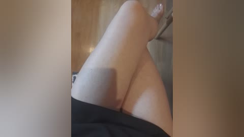 A video of a woman's legs, showing smooth skin and a black skirt pulled down to reveal her pubic area, taken in a dimly lit room with wooden walls.