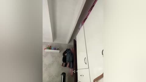 A video of a narrow, white-walled hallway with a child sitting on a gray carpet, playing with toys on a shelf. The hallway leads to a white wardrobe with a pink curtain.