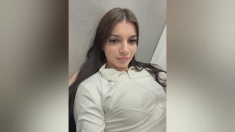 Video of a young woman with fair skin, long brown hair, wearing a white jacket, standing against a textured wall, smiling softly.