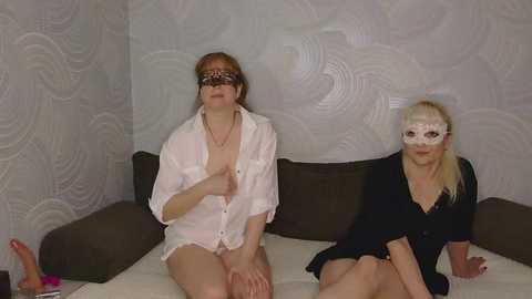 Video of two middle-aged women, one with a blindfold, wearing lingerie, sitting on a bed with a patterned wallpaper background.