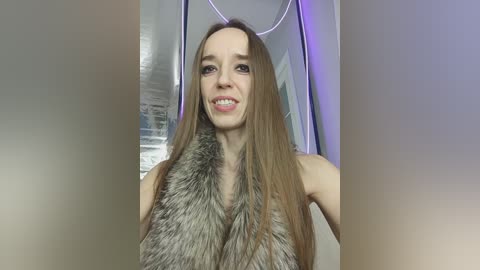 Video of a smiling, fair-skinned woman with long, straight, light brown hair, wearing a gray fur vest, in a modern room with a blurred background.