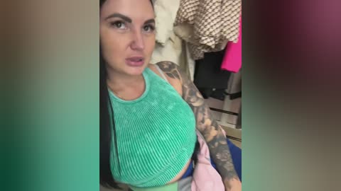 Video of a tattooed woman in a teal ribbed tank top, looking serious with a green and pink background.