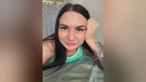 A video of a smiling woman with long black hair, wearing a green tank top and a grey jacket, lying on a bed, showcasing a tattooed arm.