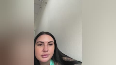 Video of a young woman with long black hair, wearing a turquoise top, standing indoors with a large, white, cylindrical object obstructing her view.