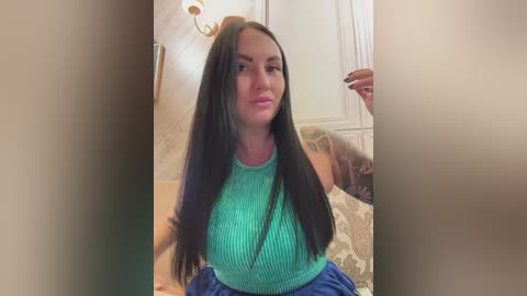 A video of a woman with long black hair, wearing a green ribbed top, sitting indoors with beige walls and a patterned couch in the background.