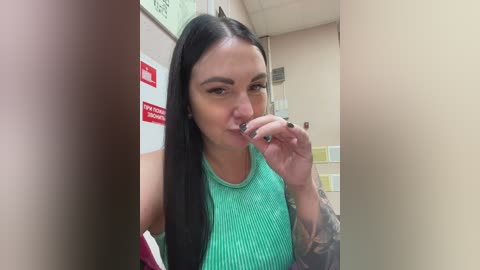 Video of a tattooed woman with long black hair, wearing a turquoise ribbed tank top, brushing her teeth in a public restroom. Background shows tiled walls, a red sign, and a yellow towel.