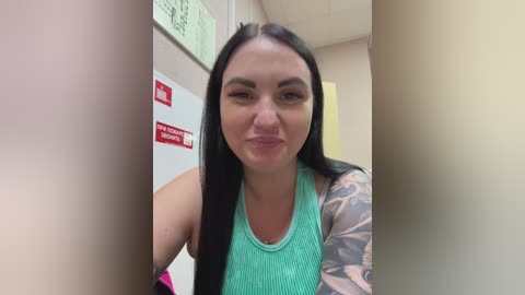 Video of a young woman with long black hair, light skin, and a tattooed arm, wearing a turquoise tank top, standing in a hallway with red signs and green walls.