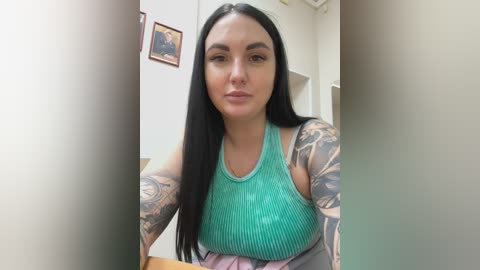 Video of a woman with long black hair, fair skin, and numerous tattoos on her arms, wearing a green ribbed tank top and a pink skirt, standing indoors with beige walls and framed art.