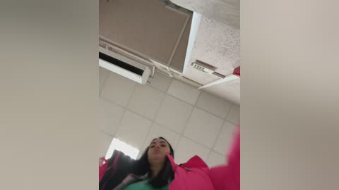 Video of a young woman with long dark hair, wearing a bright pink jacket, standing in a dimly lit room with white tiles and an air conditioning vent.