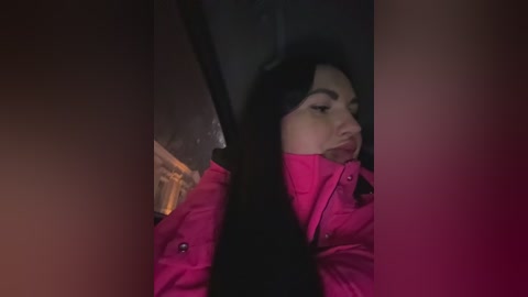 A dimly lit video shows a young woman with long black hair and fair skin, wearing a bright pink jacket, looking downcast. The background is blurred, possibly inside a vehicle.