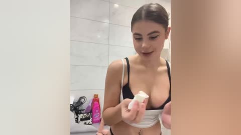 Video of a young woman with medium skin tone and brown hair tied back, wearing a black and white sports bra, applying cream to her breasts in a modern, white-tiled bathroom.