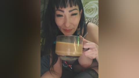 A video of an Asian woman with long black hair and light skin, wearing a black tank top, drinking coffee from a glass cup, with a floral patterned background.