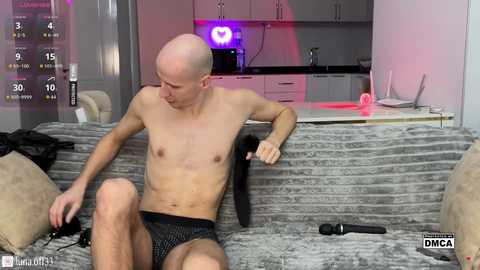 Video of a shirtless, slender, bald man with fair skin, wearing black briefs, seated on a gray-patterned couch, holding a black dildo. Background shows a modern kitchen with white cabinets and purple lighting.