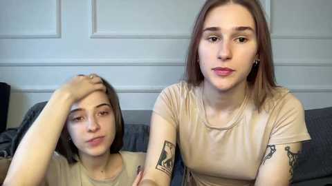 Video of two young women with light skin, one with short brown hair, another with shoulder-length brown hair. Both wear beige t-shirts; one resting her head on her hand, the other looking pensive.