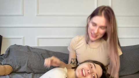 Video of two young women, one in a beige top, lying on a gray bedspread, laughing joyfully. The background features a white-paneled door.