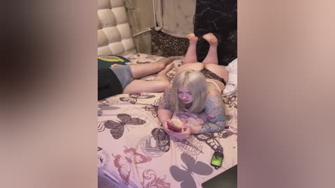 Video of a woman with long blonde hair, inked arms, and black lingerie, lying on a bed with a patterned duvet, holding a phone, in a dimly lit bedroom.