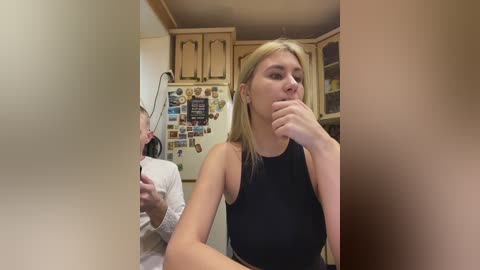 A video of a blonde woman with fair skin and a sleeveless black top, biting her finger in a kitchen with wooden cabinets and a cluttered fridge.