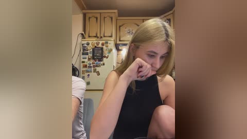 Video of a blonde woman in a black sleeveless top, sitting with her head in her hands, in a cluttered, beige kitchen with wooden cabinets and a refrigerator covered in magnets.