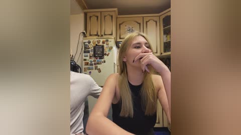 Video of a blonde woman in a black top, seated in a kitchen, resting her chin on her hand, with a fridge adorned with magnets in the background.