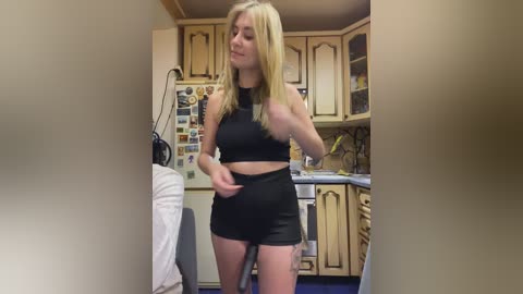 Video of a blonde woman in a black crop top and shorts, standing in a kitchen with wooden cabinets, a fridge, and a counter, holding a towel.