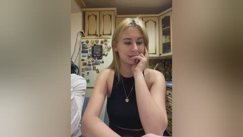 Video of a young blonde woman in a black crop top, sitting in a cozy kitchen with wooden cabinets, a fridge adorned with magnets, and a white counter.