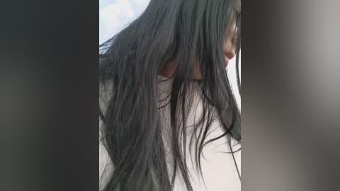 Video of a woman with long, wet black hair cascading down her back, partially obscuring her face, wearing a white top. Background is blurred, focusing on the hair texture.