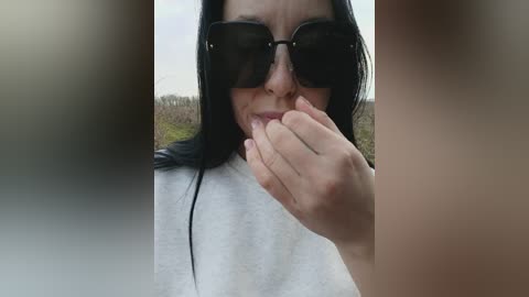 A video of a woman with long black hair and dark sunglasses, covering her mouth with her hand, standing outdoors in a blurred, grassy background.