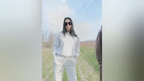 Video of a woman with long black hair, wearing white hoodie and pants, standing on a dirt path in a grassy field under a partly cloudy sky.