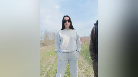 Video of a woman with long black hair, wearing sunglasses, a white hoodie, and pants, standing on a grassy path with trees and a cloudy sky in the background.