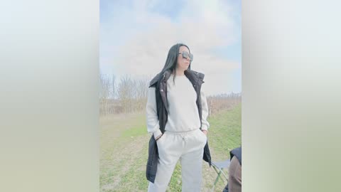 A video of a slim, long-haired woman in a white hoodie and pants, sunglasses, standing confidently on a grassy path. She holds a bike, with a blurred background of trees and a cloudy sky.