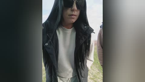 Video of a person with long black hair, sunglasses, black vest over white t-shirt, standing outdoors near a grassy area with blurred background.
