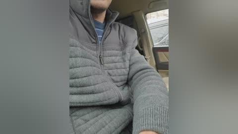Video of a person wearing a gray, quilted jacket with a blue shirt inside, driving a car with beige seats and a visible window.