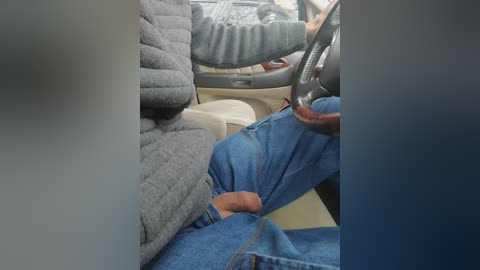 Video of a person in blue jeans and a gray jacket sitting in a car, steering wheel in hand, with a visible erect penis. The background shows a beige interior and a partially visible driver's seat.