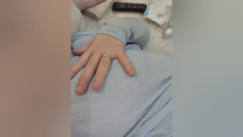 A video showing a light-skinned hand with a ring resting on a pale blue shirt, set against a blurred background with a remote control and a tissue box.