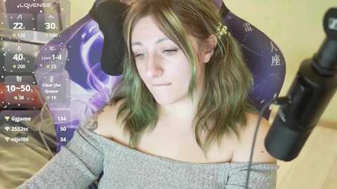 Video of a light-skinned woman with green hair in pigtails, wearing a grey off-shoulder sweater, sitting in a purple gaming chair, with a black microphone in front.