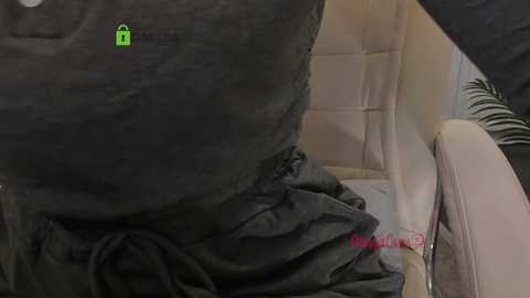 A video of a person wearing a dark gray, long-sleeved shirt with a green heart symbol on the chest, sitting on a light beige cushioned chair in a modern, neutral-toned room.