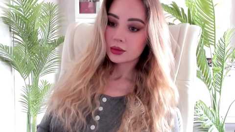 Video of a young woman with long, wavy blonde hair, fair skin, and full makeup, sitting in a white chair, surrounded by green plants. She wears a dark gray, button-up top.