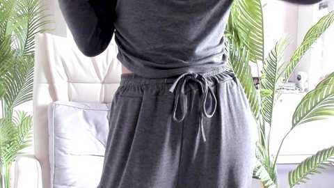 Video of a person wearing a grey, long-sleeved hoodie and matching grey sweatpants, standing in a bright, indoor room with white furniture and green potted plants in the background.