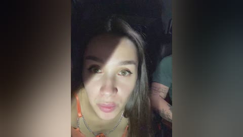 Video of a young woman with long brown hair, light skin, and green eyes, wearing an orange top, sitting indoors, partially obscured by shadow.