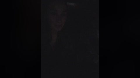 A dimly lit, grainy video of a young woman with long, dark hair, partially obscured by shadows, wearing a dark jacket, standing in a dark room.
