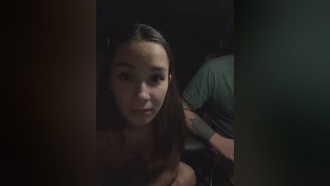 A dimly-lit video of a young woman with long brown hair, wearing a dark top, sitting on a black car seat, partially obscured by shadows.