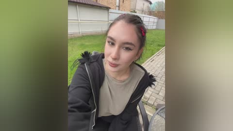 Video of a young Asian woman with dark hair styled in a side ponytail, wearing a black jacket, standing in a suburban backyard with a brick pathway and green grass.