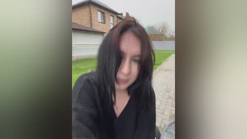 A video of a young woman with straight, shoulder-length dark brown hair, wearing a black hoodie, standing on a paved pathway beside a suburban house with a garage.